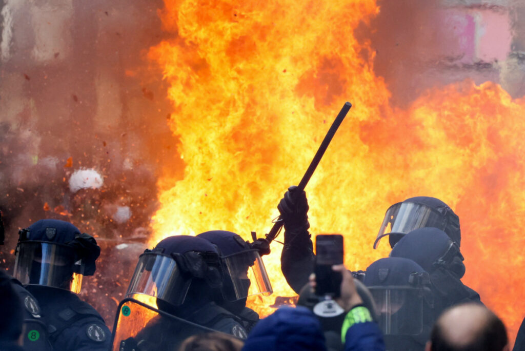 Flames from a fire rise behind law enforcement officers during a demonstration against the French government's pension reform plan as part of the fourth day of national protests, in Paris, France, February 11, 2023.