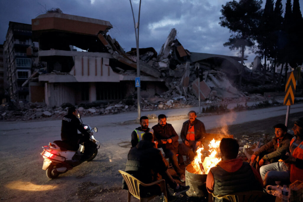 People warm themselves by a fire beside a collapsed building and rubble, in the aftermath of a deadly earthquake, in Antakya, Hatay province, Turkey, February 21, 2023.