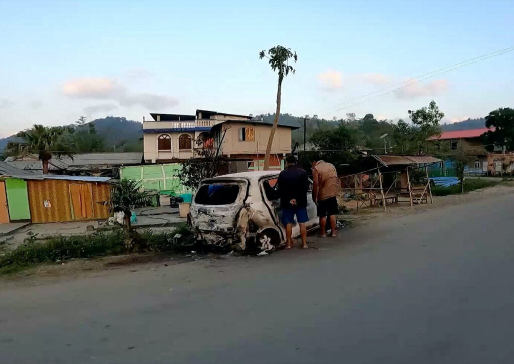 People stand near a burnt vehicle, in Manipur, India, on 6th May, 2023, in this screengrab obtained from a social media video.