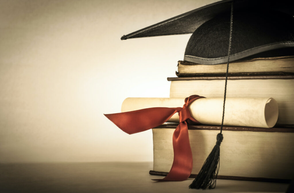 A mortarboard and graduation scroll, tied with red ribbon, on a stack of old battered book with empty space to the left.