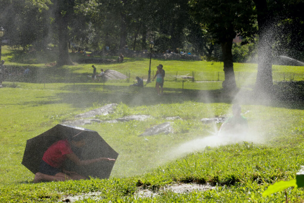 A child takes shelter under an umbrella while another one plays with a water sprinkler, in Manhattan's Central Park, in New York City, US, on 28th July, 2023.