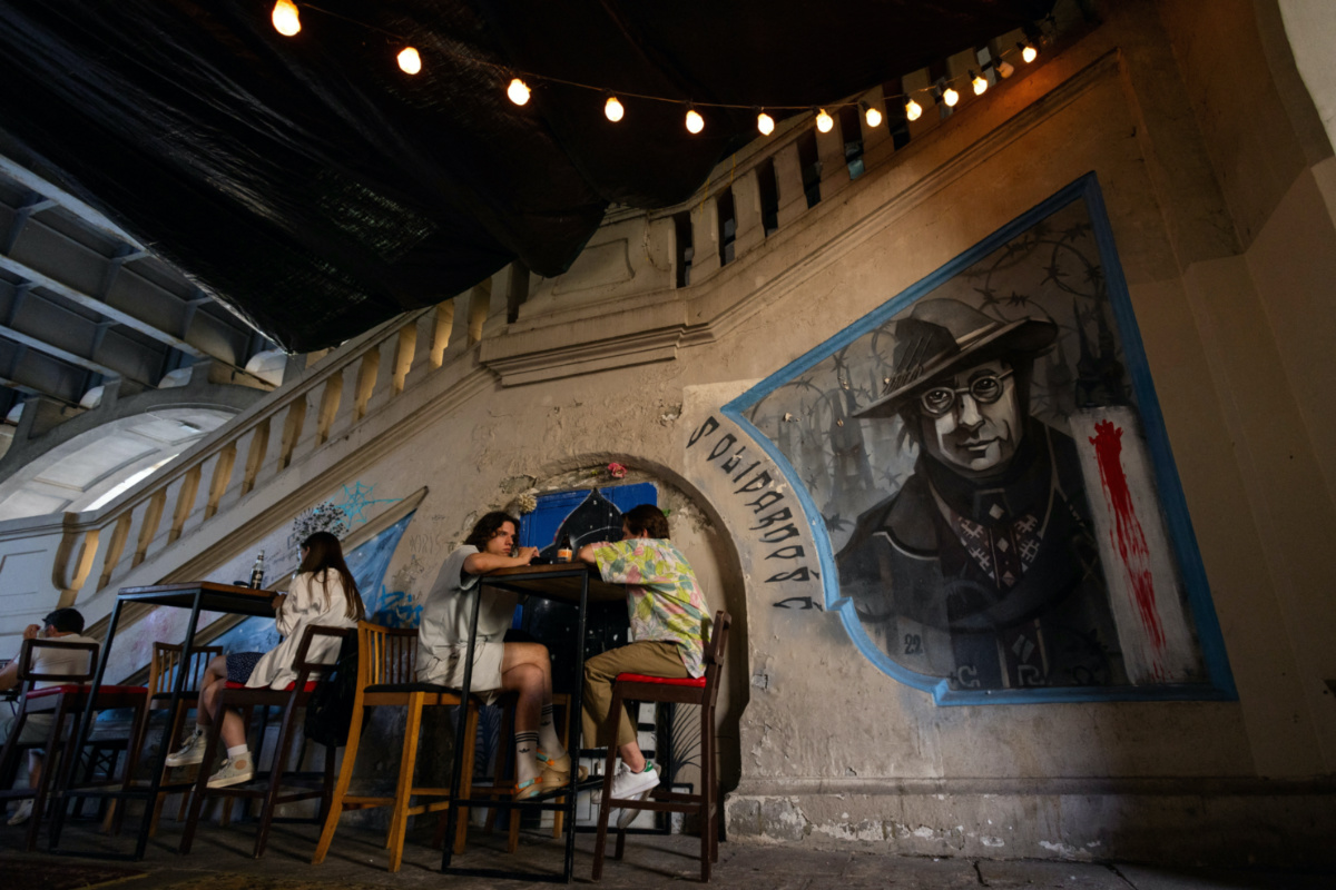People sit at a Belarusian bar next to a graffiti commemorating Ales Pushkin, a Belarusian artist and political prisoner who died this year in Belarusian jail under unexplained circumstances, in Warsaw, Poland, on 18th July, 2023