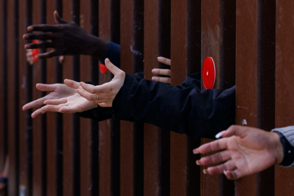 Migrants reach out from the border wall for food from an aid agency as the wait between the primary and secondary border fences.