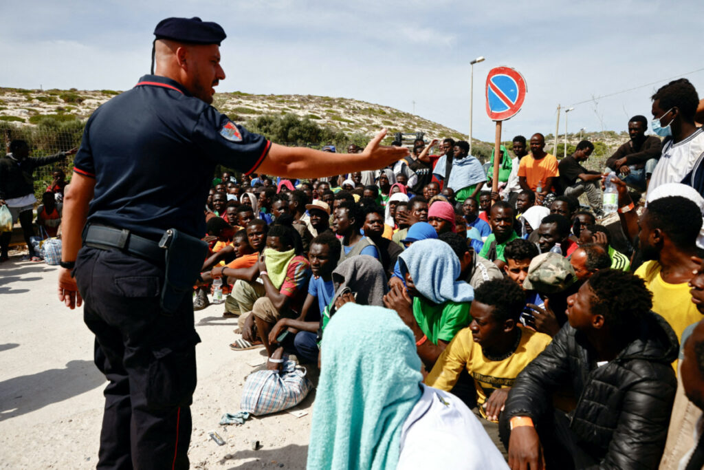 A member of the Carabinieri gestures towards migrants outside the hotspot, on the Sicilian island of Lampedusa, Italy, on 16th September, 2023