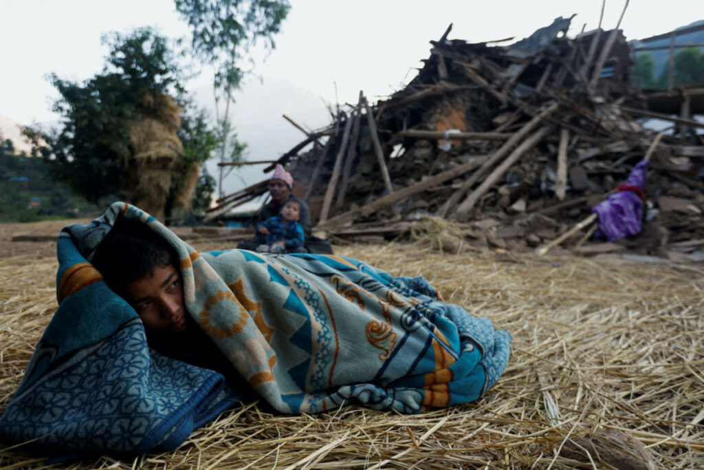 A boy covers himself with a blanket as he lies down on a floor next to a house collapsed during an earthquake in Jajarkot, Nepal, on 5th November, 2023