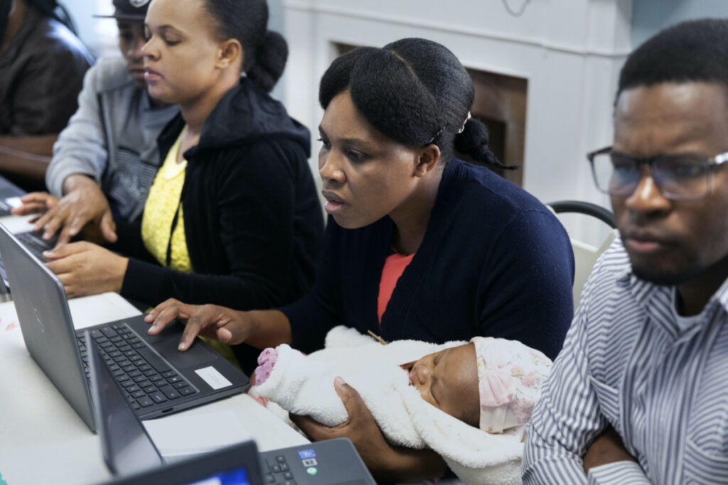 Jimene Admettre, center, sits next to her husband Ernseau, right, and holds her daughter Gabyana while learning computer skills, on Friday, 22nd December, 2023, in a rectory building where they are staying at the Bethel AME Church in the Jamaica Plain neighbourhood of Boston.