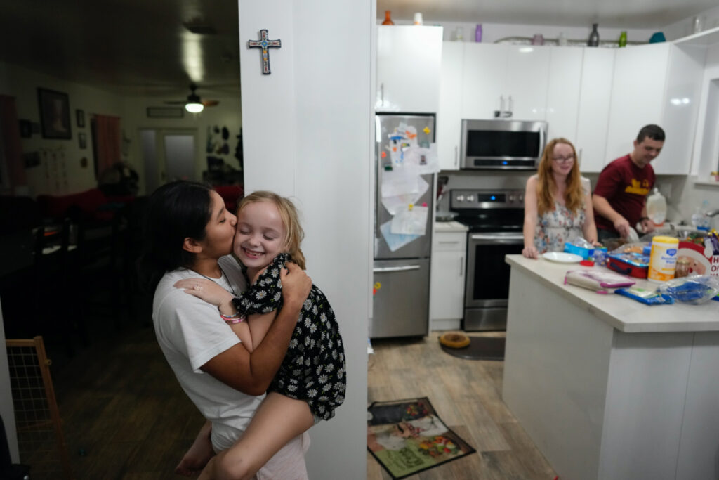 Sol, left, a 14-year-old from Argentina, kisses eight-year-old Maddie Hazelton as they play together in the kitchen of Sol's foster parents, Andy, right, and Caroline Hazelton, in Homestead, Florida, on Monday, 18th December, 2023.